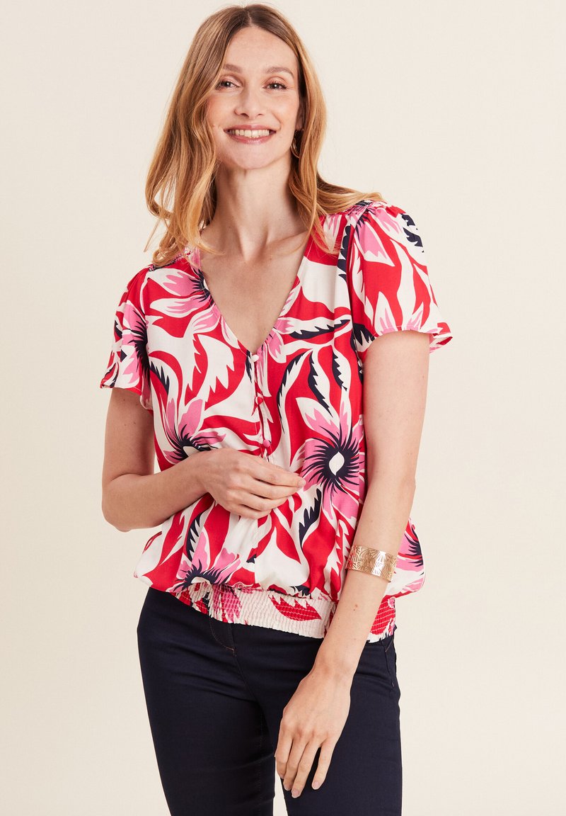 Bluse mit Blumenmuster in Rot, Pink und Schwarz, V-Ausschnitt, kurzen Ärmeln und gerafftem Bund. Kombiniert mit einer dunklen Hose.