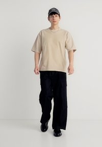 Νεαρός άνδρας που περπατά μπροστά φορώντας μια μπεζ φαρδιά μπλούζα, μαύρο παντελόνι καρό, μαύρα παπούτσια και σκούφο μπέιζμπολ σε απλό λευκό φόντο.