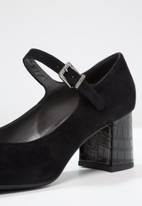 Chaussure Mary Jane en daim noir avec un talon texturé en croûte de crocodile noir. Dotée d'une bride de cheville réglable avec une boucle métallique.