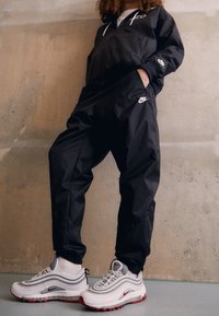 Svart Nike vindjacka med elastiska muddar, kombinerad med svarta joggingbyxor. Fötterna bär grå och vita Nike Air Max 97-skor med röda detaljer.