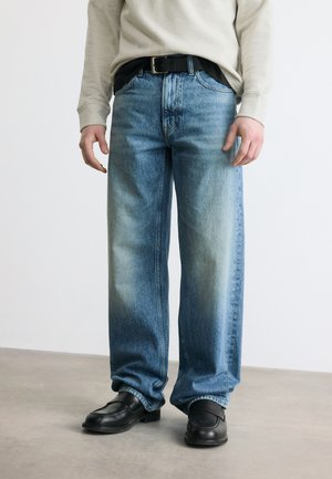 Hombre con vaqueros azules desteñidos de corte holgado, cinturón negro, sudadera beige y zapatos de cuero negros, de pie sobre un suelo gris.