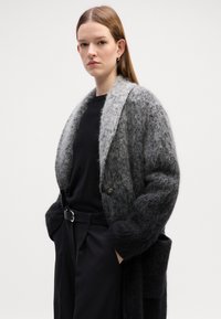 Cappotto lungo in misto lana con sfumature di grigio a gradiente, colletto a scialle, chiusura con un bottone e tasche frontali. Indossato sopra una maglia nera.