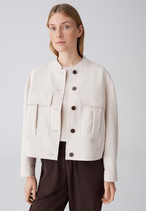 Vrouw die een crème kleurige jas met grote zakken aan de voorkant en donkere bruine broek draagt, staand tegen een effen lichte achtergrond.