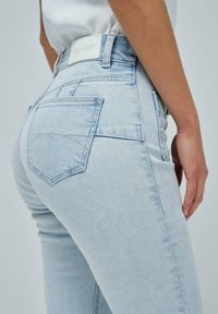 Jeans de mezclilla en azul claro con un diseño ajustado, que presentan un bolsillo trasero y una etiqueta de logo blanco en la cintura. Textura suave, corte clásico.