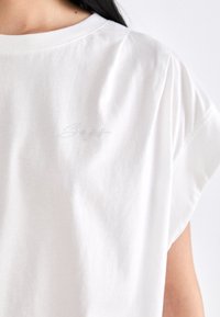 T-shirt bianca in cotone con maniche corte, caratterizzata da un sottile design di un logo con borchie argentate nella zona sinistra del petto. Texture morbida.