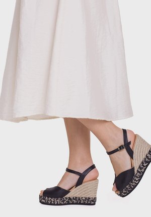 Nogi w czarnych skórzanych sandałach na koturnie z paskiem wokół kostki i plecionymi obcasami espadrylowymi, zestawione z białą, zwiewną spódnicą o długości do połowy uda.