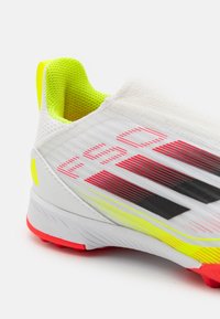 Scarpa da ginnastica bianca con superficie testurizzata, caratterizzata da accenti rossi e neri, un tallone giallo fluo e una suola flessibile con tacchetti rossi.