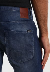 Mörkblå denimjeans med en läderbakpatch och svart etikett. Har en myntficka och förstärkt sömnad på bakfickorna.