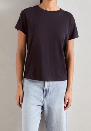 Kvinde iført en ensfarvet mørk kortærmet t-shirt, der er let stukket ned i lyseblå højttaljede jeans, stående mod en neutral baggrund.