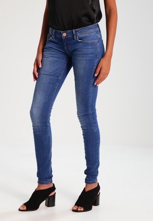 Woman wearing fitted blue jeans and black open-toe heeled sandals, standing with hands resting by her sides.