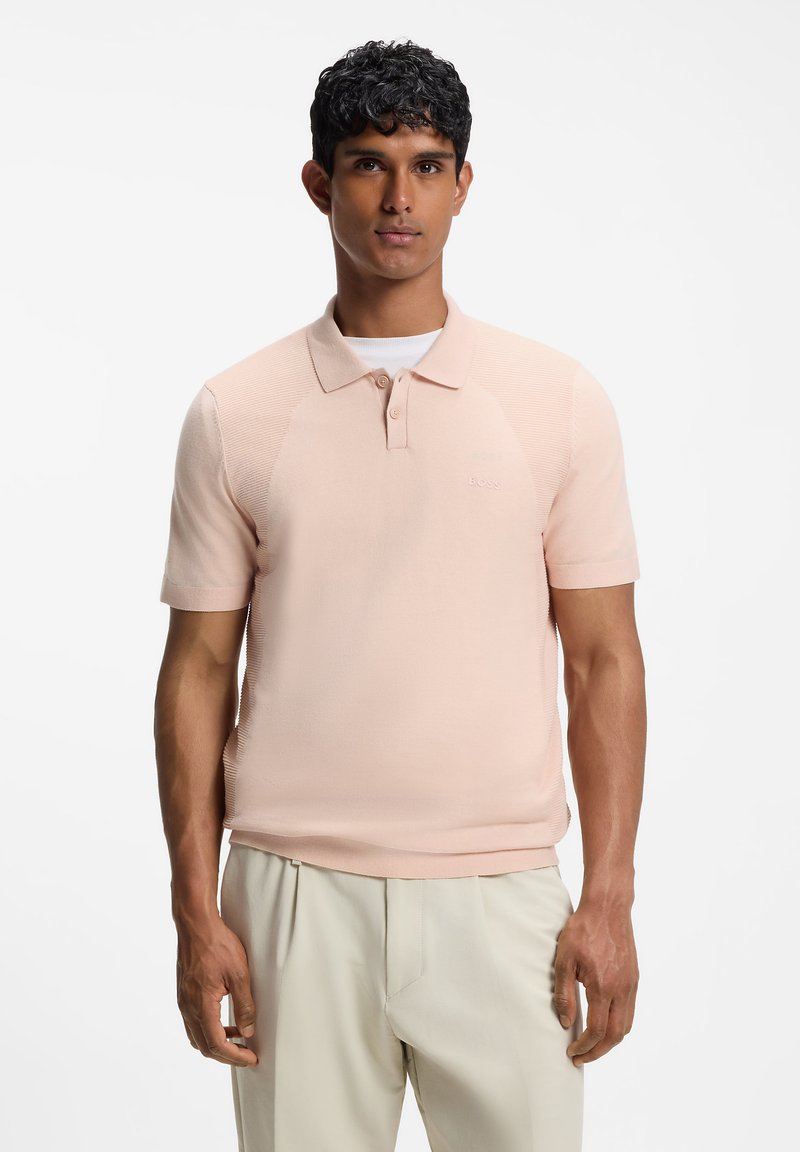 Homme aux cheveux courts et bouclés portant un polo rose clair à manches courtes et un pantalon beige, debout sur fond blanc.