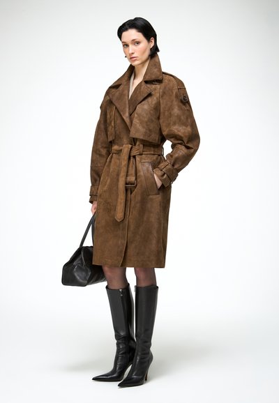 Žena v hnedom semišovom trenčkot s opaskom, čiernych čižmách nad kolená, držiaca čiernu koženú kabelku, stojaca na bielom pozadí.