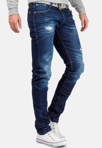Jeans in denim blu scuro con taglio slim, caratterizzati da aree scolorite e un leggero sbiadimento. Abbinati a sneaker bianche con dettagli rossi.