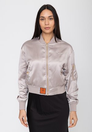 Frau mit langem dunklem Haar, die eine glänzende hellgraue Bomberjacke mit goldenem Reißverschluss und einem schwarzen Rock vor einem schlichten weißen Hintergrund trägt.