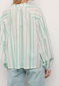 Donna che indossa una blusa lunga e larga con maniche lunghe a righe verticali verdi, blu e bianche, abbinata a jeans di denim blu chiaro, vista da dietro.