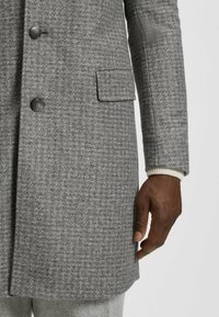 Mans hand bredvid en grå houndstooth-mönstrad kappa med svarta knappar och ficklock, vit tröja är synlig under kappans ärm.