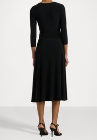 Černé šaty s přiléhavým vrškem, tříčtvrtečními rukávy a rozšířenou sukní. Hladká látka a délka do kotníků s černými sandály na vysokém podpatku.