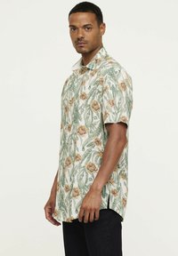 Korte mouwen shirt gemaakt van lichte stof met een bloemenpatroon dat groene bladeren en gele bloemen bevat. Zijsplitten verbeteren de luchtstroom.