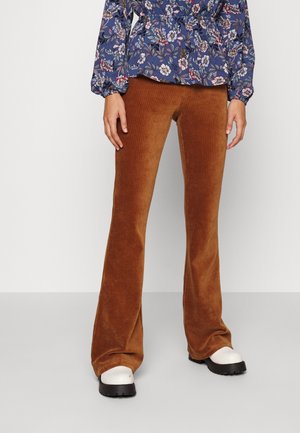 Erwachsener trägt braune Cord-Flared-Hosen, weiße Schuhe mit klobiger Sohle und ein blaues, langärmliges Oberteil mit Blumenmuster und steht vor einem schlichten Hintergrund.
