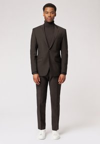 Dunkelbrauner Anzug mit Reverskragen, zwei vorderen Taschen und schmaler Passform, kombiniert mit einem schwarzen Rollkragenpullover und weißen Sneakers.