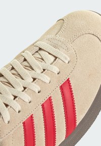 Μπεζ σουέτ αθλητικά παπούτσια με κόκκινες δερμάτινες ρίγες, υφή επιφάνειας, επίπεδα κορδόνια και καφέ λάστιχο σόλα. Λεπτομέρεια ενισχυμένου καπακιού δαχτύλων.