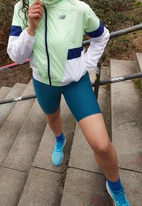 Jasnozielona i biała kurtka na zamek, niebieskie szorty i turkusowe buty sportowe. Gładka tkanina, sportowy krój, widoczne logo, stoi na betonowych schodach.