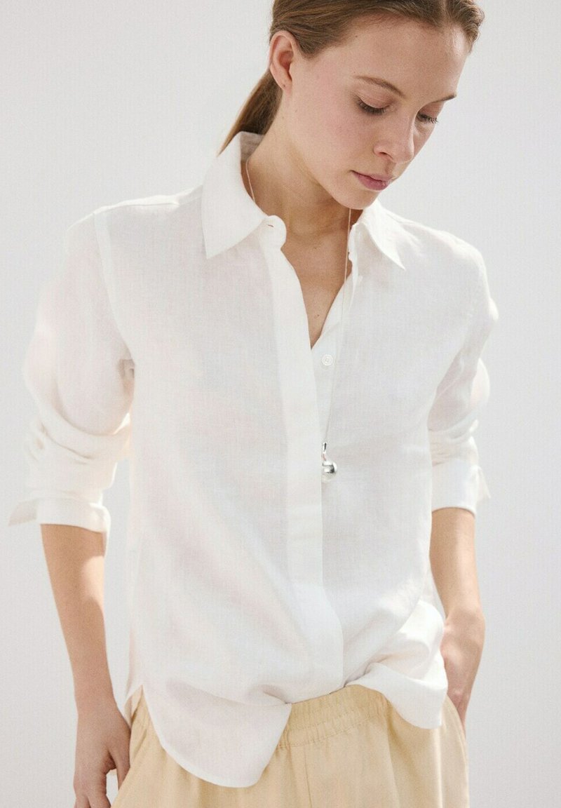 Frau trägt ein weißes, aufgeknöpftes Hemd mit hochgekrempelten Ärmeln und beige Hosen, blickt nach unten mit den Händen in den Taschen vor einem schlichten Hintergrund.