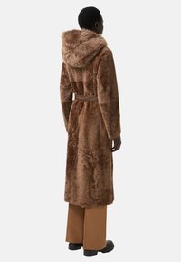 Hnědý kabát z umělé kožešiny s kapucí, opaskem v pase a dlouhými rukávy. Má měkkou texturu a střední délku.