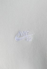 Bílá texturovaná tkanina se bílou vyšívanou logem Nike SB v dolním rohu. Čistý, jednoduchý design bez dalších vzorů.