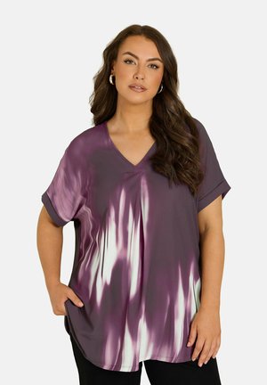 Femme aux longs cheveux ondulés portant un t-shirt tie-dye violet et blanc à col en V et un pantalon noir, debout avec une main dans sa poche.