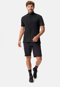 Schwarze ärmellose Radweste mit hohem Kragen, kombiniert mit schwarzen Shorts. Sie besitzt einen Reißverschluss und strukturierte Stoffakzente.
