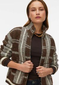 Brūns un balts rakstains kārts ar rāvējslēdzēju, izgatavots no teksturētas vilnas maisījuma, ar augstu apkakli un ribām pie aprocēm, apvienots ar tumšu augšu.