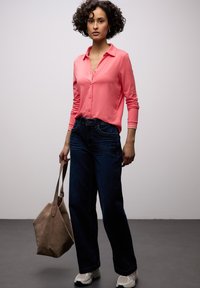 Chemise rose pâle à manches longues avec boutons, associée à un jean denim foncé à jambes larges. Porte un sac taupe doux, et des baskets blanches et grises.