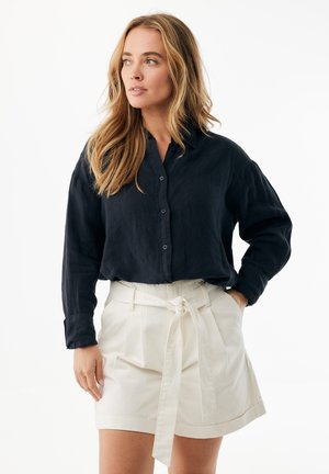 Marineblaue Hemdbluse mit langen Ärmeln, kombiniert mit weißen, hoch taillierten Shorts mit gebundenem Gürtel. Glatte Stofftexturen sind deutlich erkennbar.