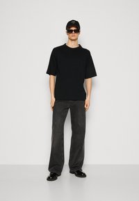 T-shirt nera oversize in cotone, abbinata a jeans a gamba larga grigio scuro. Gli accessori includono occhiali da sole neri e un cappellino nero con testo bianco.