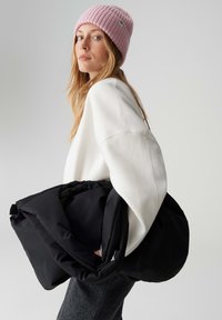 Borsa imbottita nera tenuta sotto il braccio, donna con maglione bianco oversize e berretto rosa a coste, con lunghi capelli biondi. Pantaloni grigi.