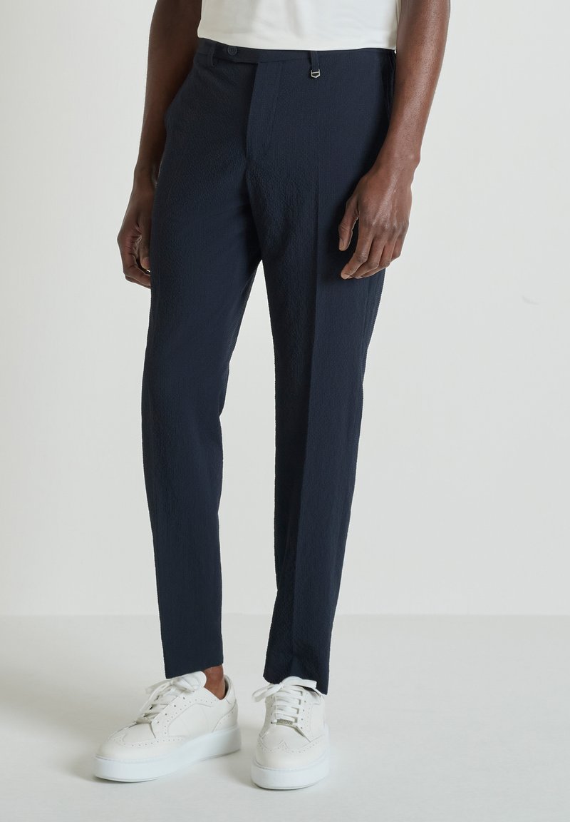 Hombre vestido con pantalones ajustados azul marino oscuro y zapatillas blancas, posando frente a un fondo claro y liso.