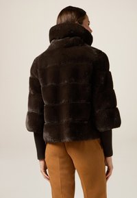 Brązowa kurtka z sztucznego futra z krótkim rękawem i szerokimi rękawami, z poziomymi, żeberkowymi wzorami oraz wysokim kołnierzem. W zestawie z beżowymi spodniami.