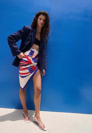 Mujer con el pelo largo y rizado, vestida con un blazer azul marino, top corto negro, falda cruzada abstracta en rojo, blanco y azul, y sandalias blancas con tacón, de pie frente a una pared azul.