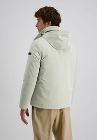 Man in een lichtgrijze hoodie en beige broek, staande zijwaarts met het hoofd naar links gekeerd tegen een effen achtergrond.