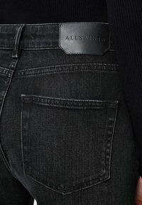 Jean en denim noir avec une texture lisse, un design classique à cinq poches, et une poche arrière cousue ornée d'un patch logo en cuir noir.