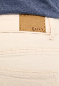 Jean en denim beige avec un patch en cuir marron clair portant l'inscription "ROXY" sur la ceinture arrière, présentant des coutures discrètes et un détail de poche.