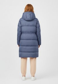 Gewatteerde jas in marineblauw met capuchon en elastische taille. De doorgestikte textuur heeft horizontale delen, gecombineerd met lichtgekleurde broek.