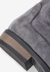 Stivaletto alla caviglia in suede grigio con una finitura testurizzata, caratterizzato da un tacco marrone impilato e una base nera liscia. Dettagli cuciti lungo le cuciture.