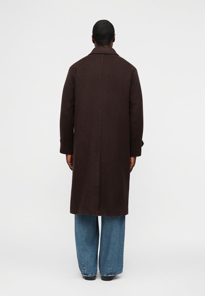 Mann von hinten gesehen, trägt einen langen dunkelbraunen Mantel über blauen Jeans und schwarzen Schuhen vor einem schlichten weißen Hintergrund.