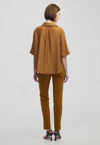 Bruin kleurige top met korte mouwen, gecombineerd met mosterdkleurige slim fit broek en zwarte gespenhakken, gezien vanuit de achterkant.