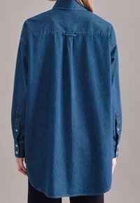 Camicia di denim blu scuro, con bottoni e colletto a punta, maniche lunghe e orlo curvo. Presenta dettagli sulla spalla posteriore e polsini con bottoni.