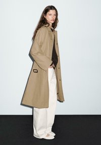 Femme aux longs cheveux ondulés portant un trench-coat beige, un haut sombre, un pantalon large blanc et des chaussures plates blanches, se tenant de profil sur un sol noir.