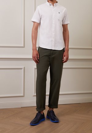 Chemise blanche à manches courtes avec boutons, pantalon tapered vert olive, et chaussures bleu foncé avec semelles bleu vif. Aucune motif ou texture visible.