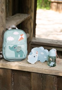 Mochila azul para niños, contenedores con forma de pájaro y botella de agua con diseños de elefante y cometa sobre superficie de madera al aire libre.